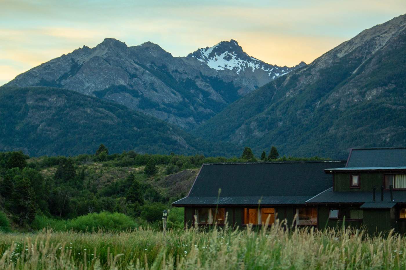 A lodge's dark-timbered exterior glowing with lit windows at dusk, mountains rising behind a foreground of tall grass