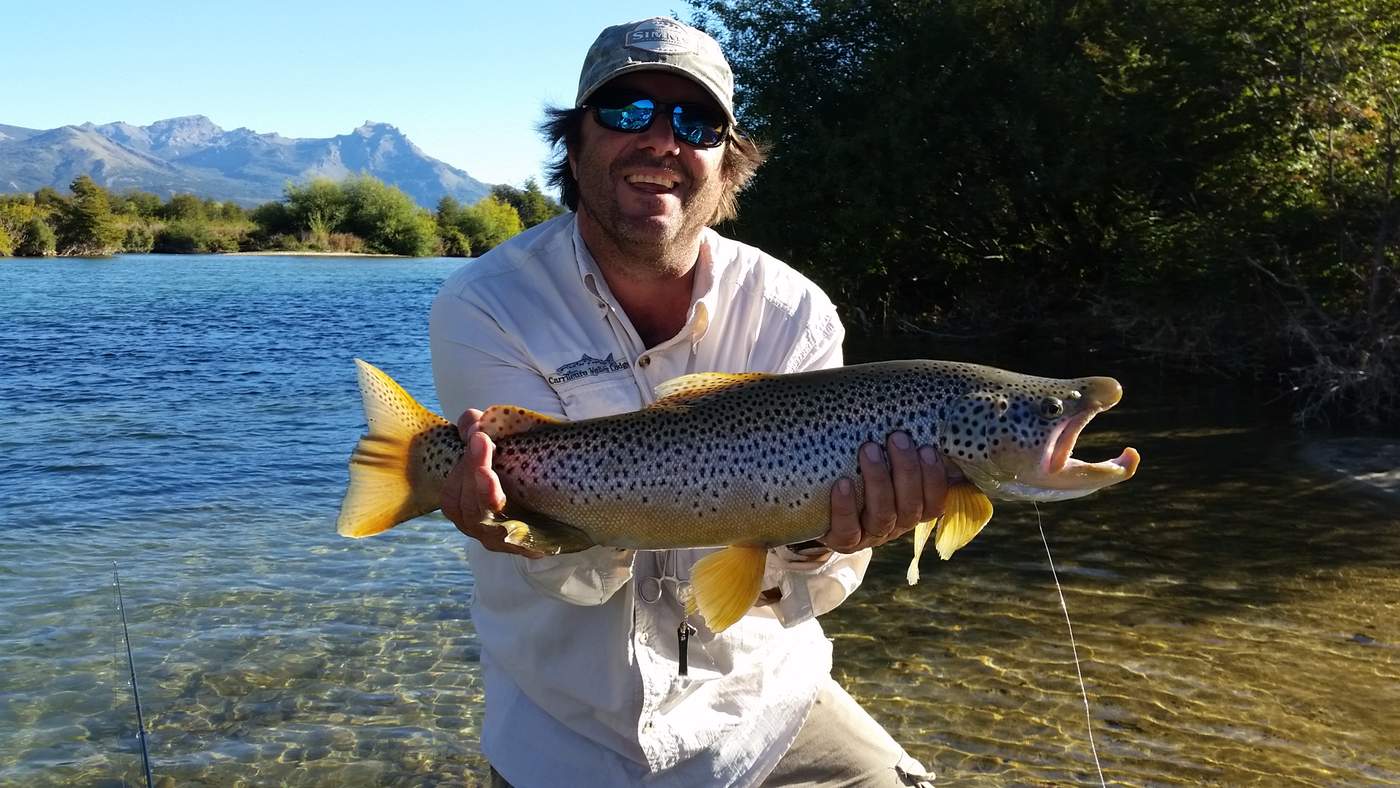 An angler holding up a large brown trout, the river and mountains behind