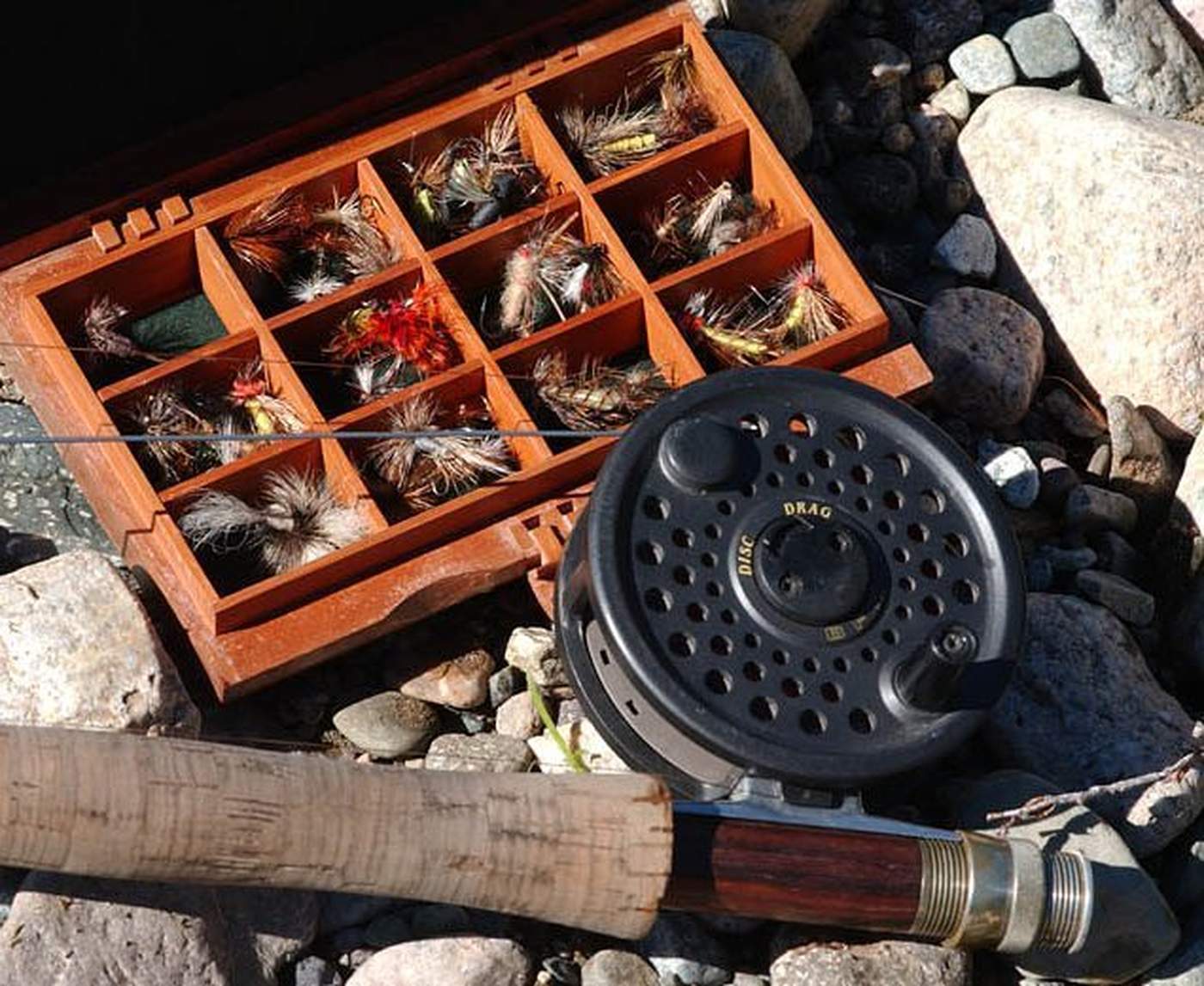 A fly box full of hand-tied flies beside a reel and cork-handled rod, resting on river stones