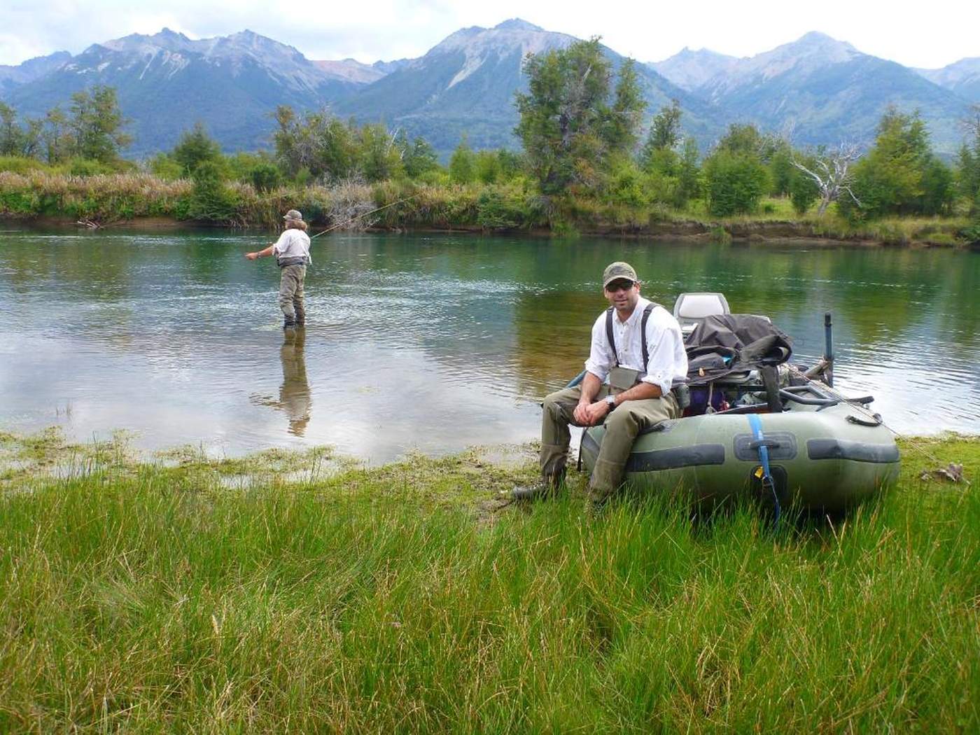 Two anglers on a grassy riverbank, one fishing and one seated by an inflatable raft, mountains beyond