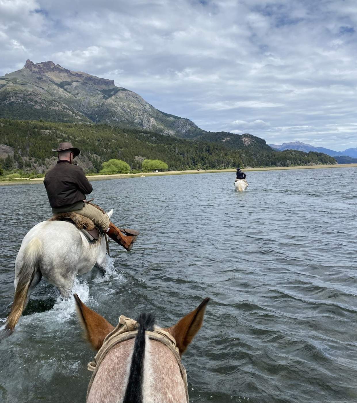 A view from the saddle, two riders crossing a lake ahead toward a jagged mountain peak