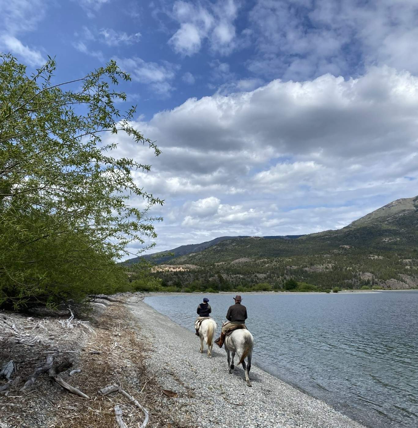Two riders on horseback along a gravel lakeshore, mountains reflected in the still water