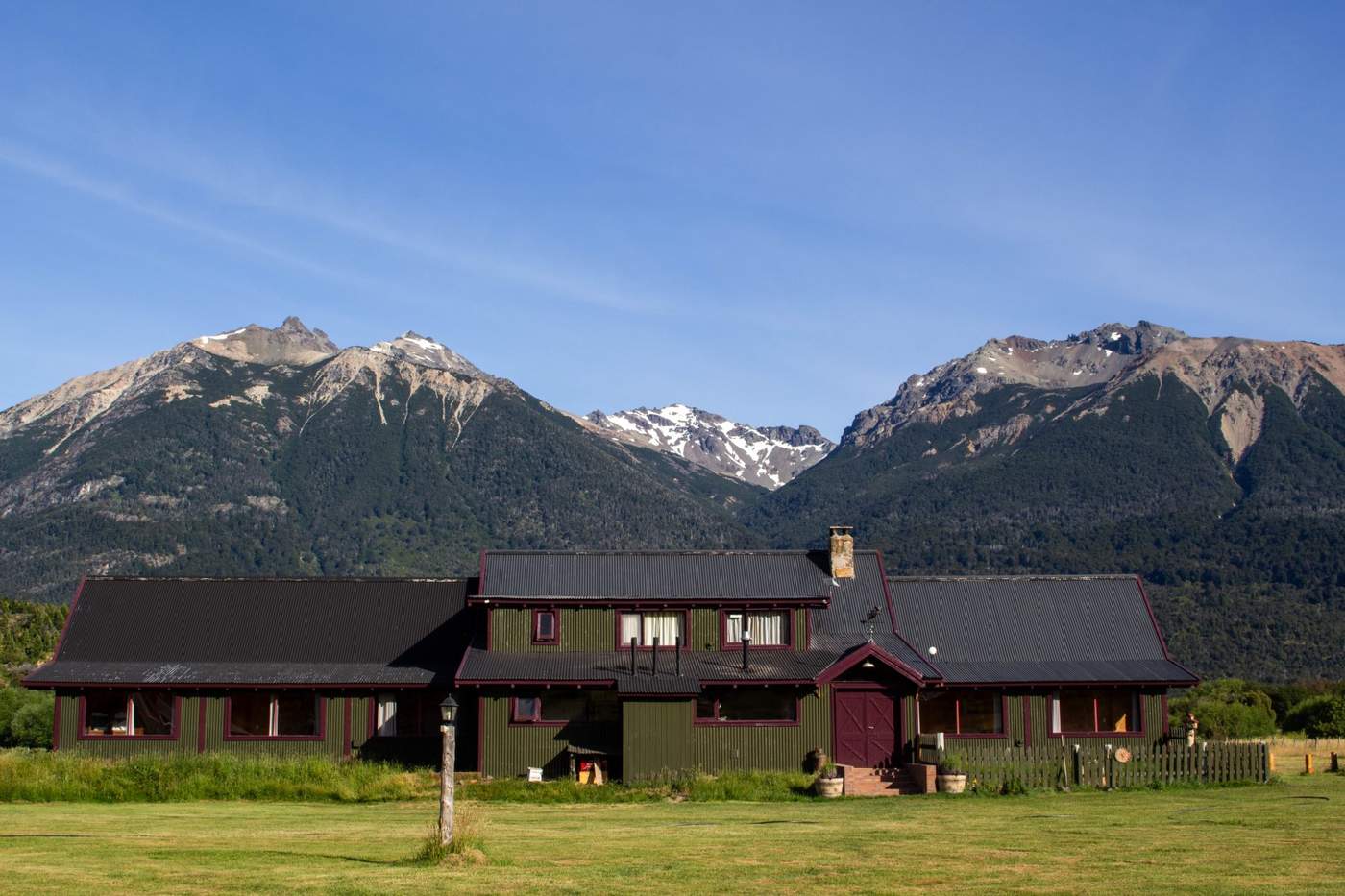 A green-and-burgundy lodge exterior beneath a steep black metal roof, snow-capped peaks above a manicured lawn