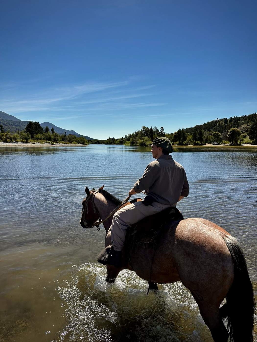 A rider on horseback fording a river, forest and mountains lining the far bank