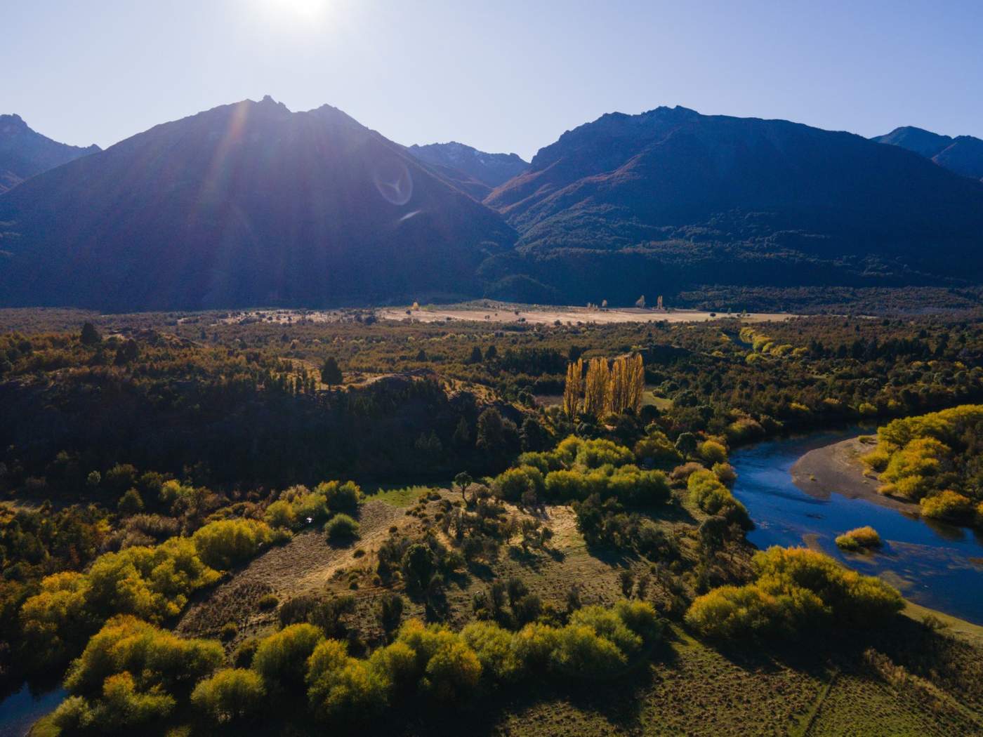 An aerial view of a valley where a river bends past golden poplars, mountains rising on either side