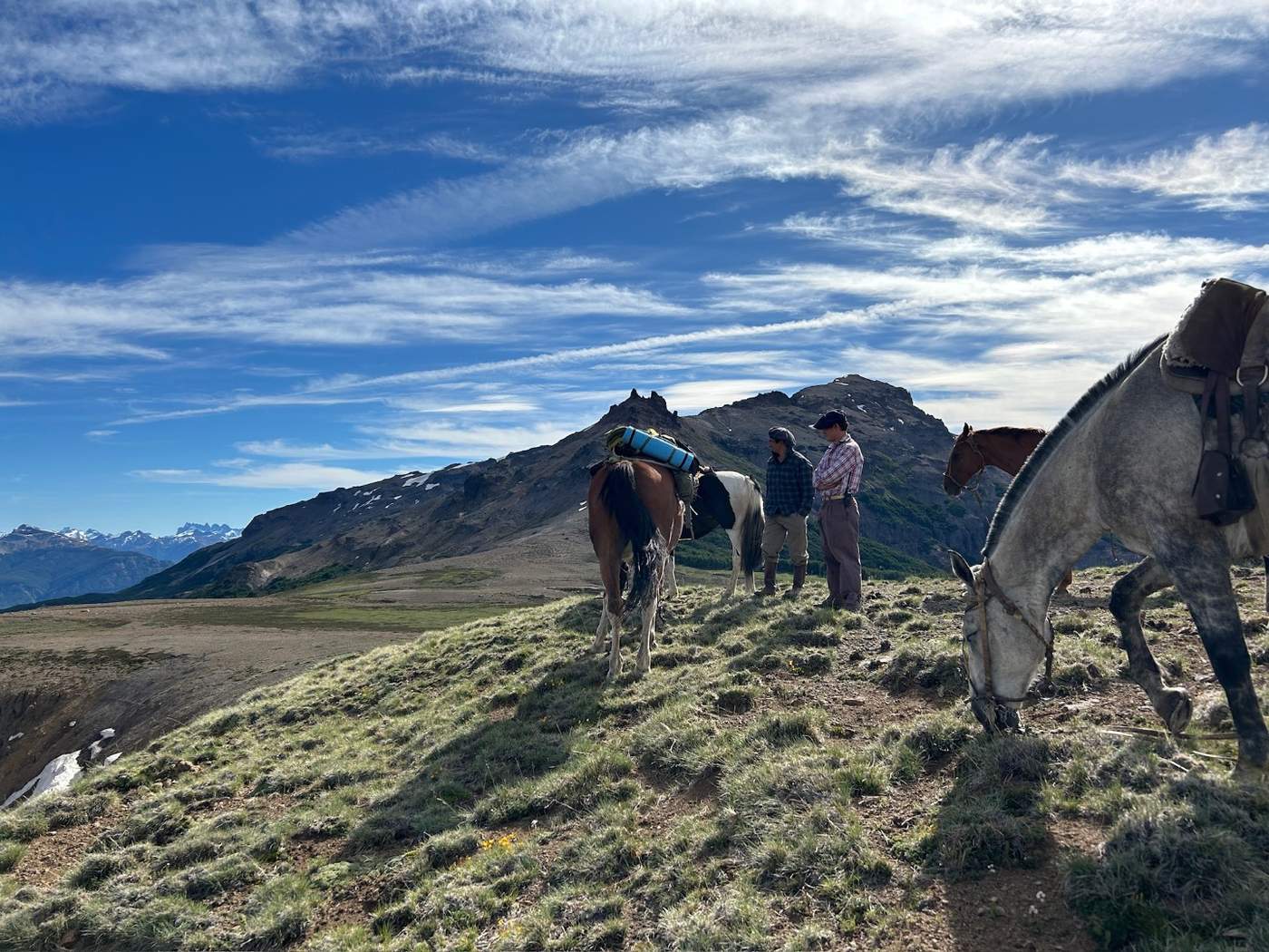 Two riders pausing on a windswept ridge with their horses, a rugged peak and distant mountain ranges behind them