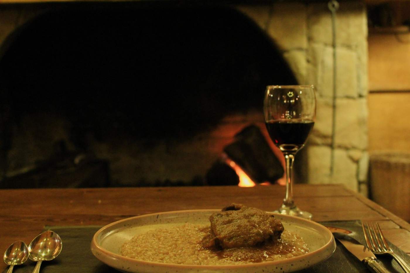 A plated dish of braised meat over risotto, a glass of red wine on the table before a glowing wood-fired oven