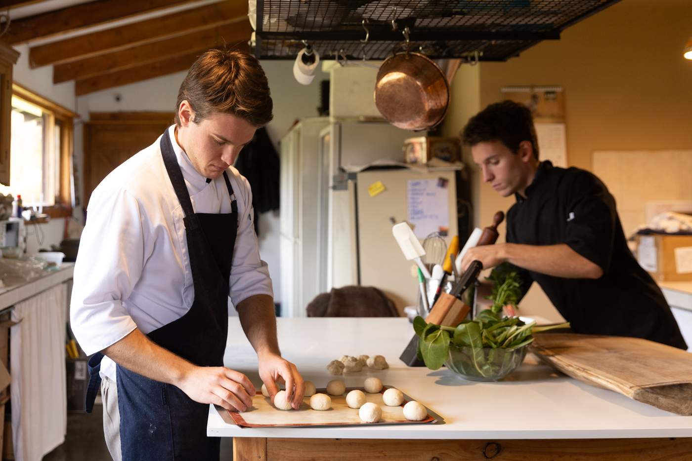 Two of the lodge's chefs at work in the kitchen, one shaping dough on a tray, the other prepping greens beneath a hanging copper pot