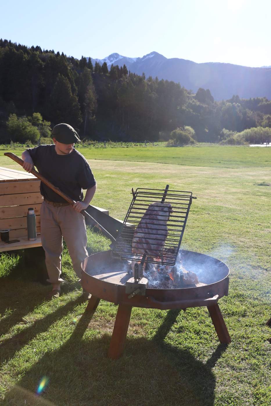 Ribs roasting over an open fire pit on the lawn, tended by a staff member, mountains behind