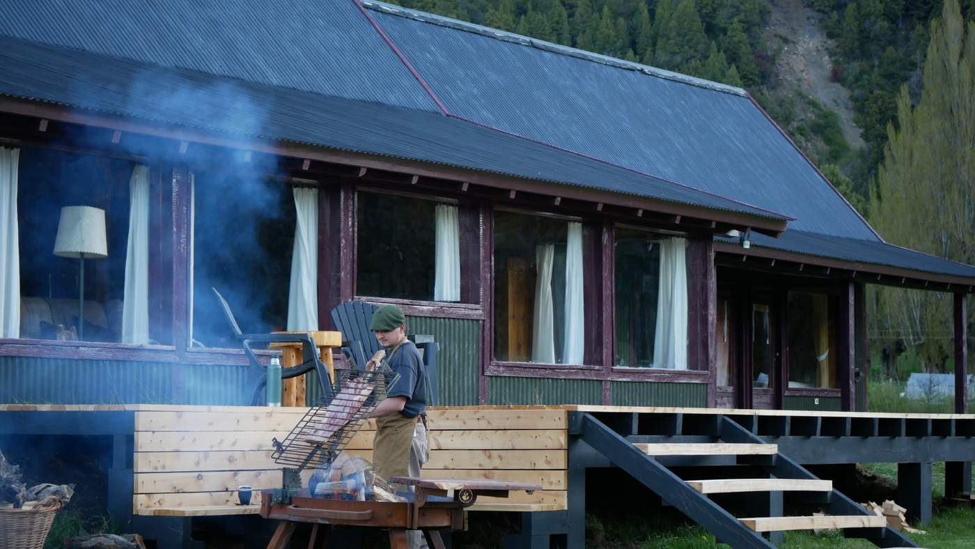 A rack of meat roasting on the lodge's back deck at dusk, a staff member tending it as smoke rises against the mountain backdrop