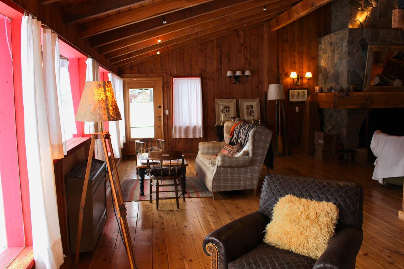 A sitting room with a plaid armchair and a wool-draped leather chair beneath a vaulted wood-beam ceiling, a stone fireplace in the corner