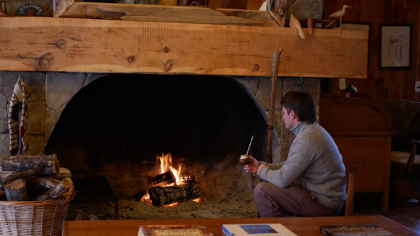 A guest seated by a crackling stone fireplace, sipping mate from a gourd beside a mantel of driftwood and carved birds