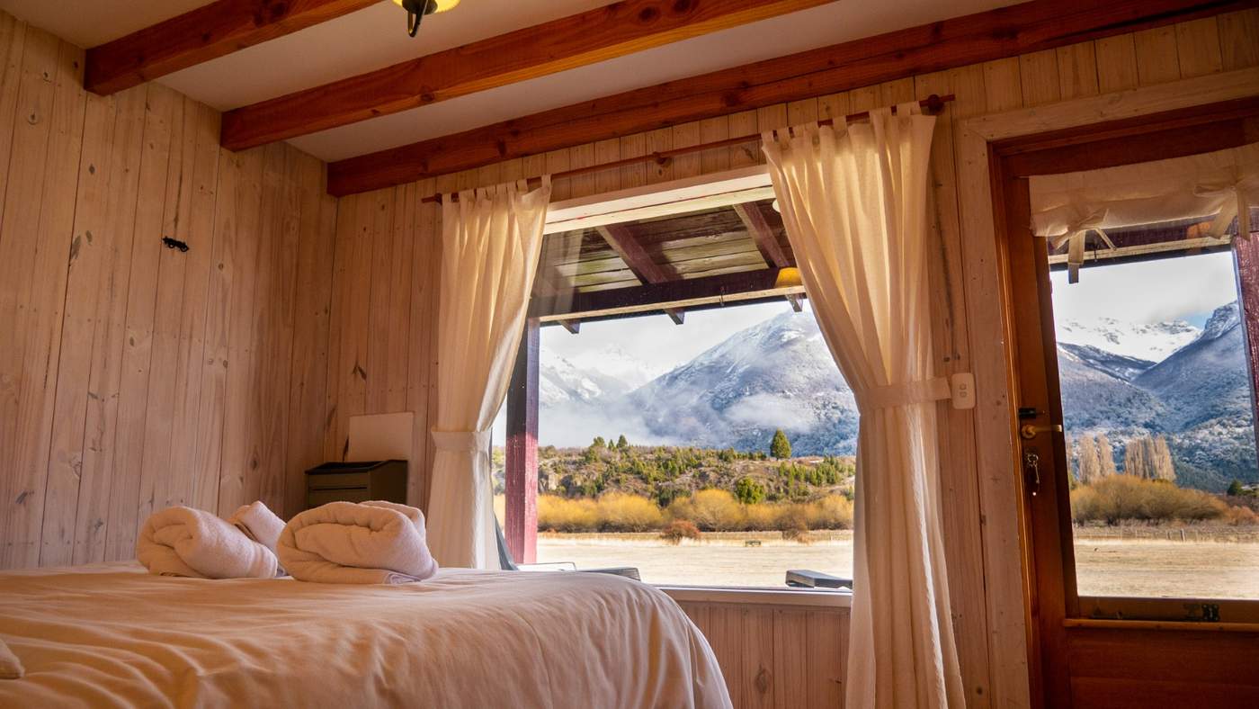 A guest room with a turned-down bed and stacked white towels, a wall of windows opening onto a mountain view
