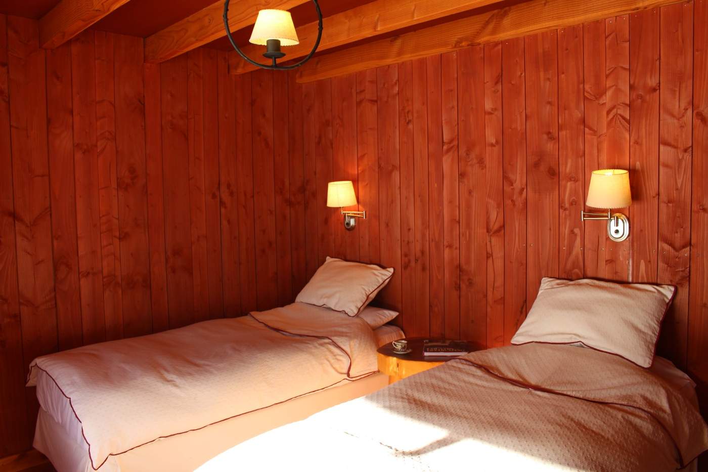 A twin-bed guest room with burgundy-trimmed white linens and wood-paneled walls, lit by wall sconces