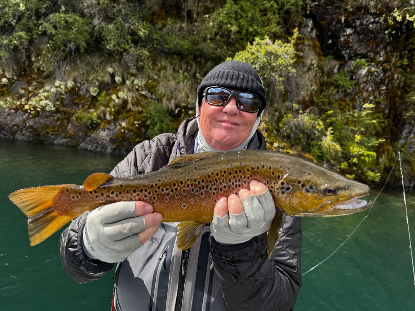A guest holding up a brown trout on a riverbank, a mossy rock face behind