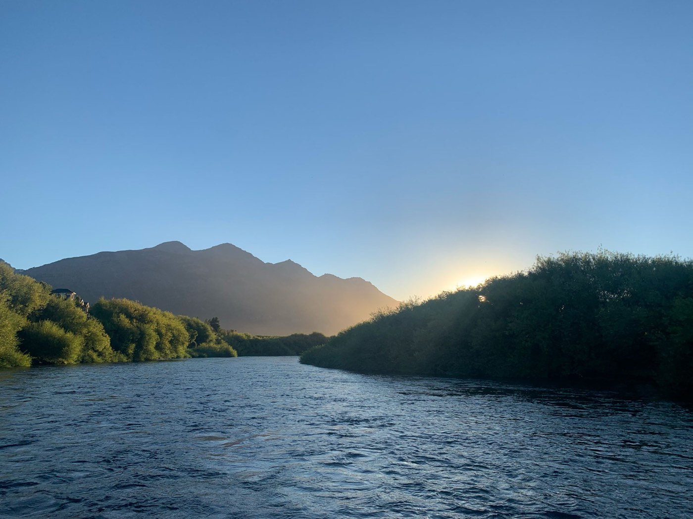 A calm river at sunrise, sunlight breaking through trees on the far bank, mountains beyond