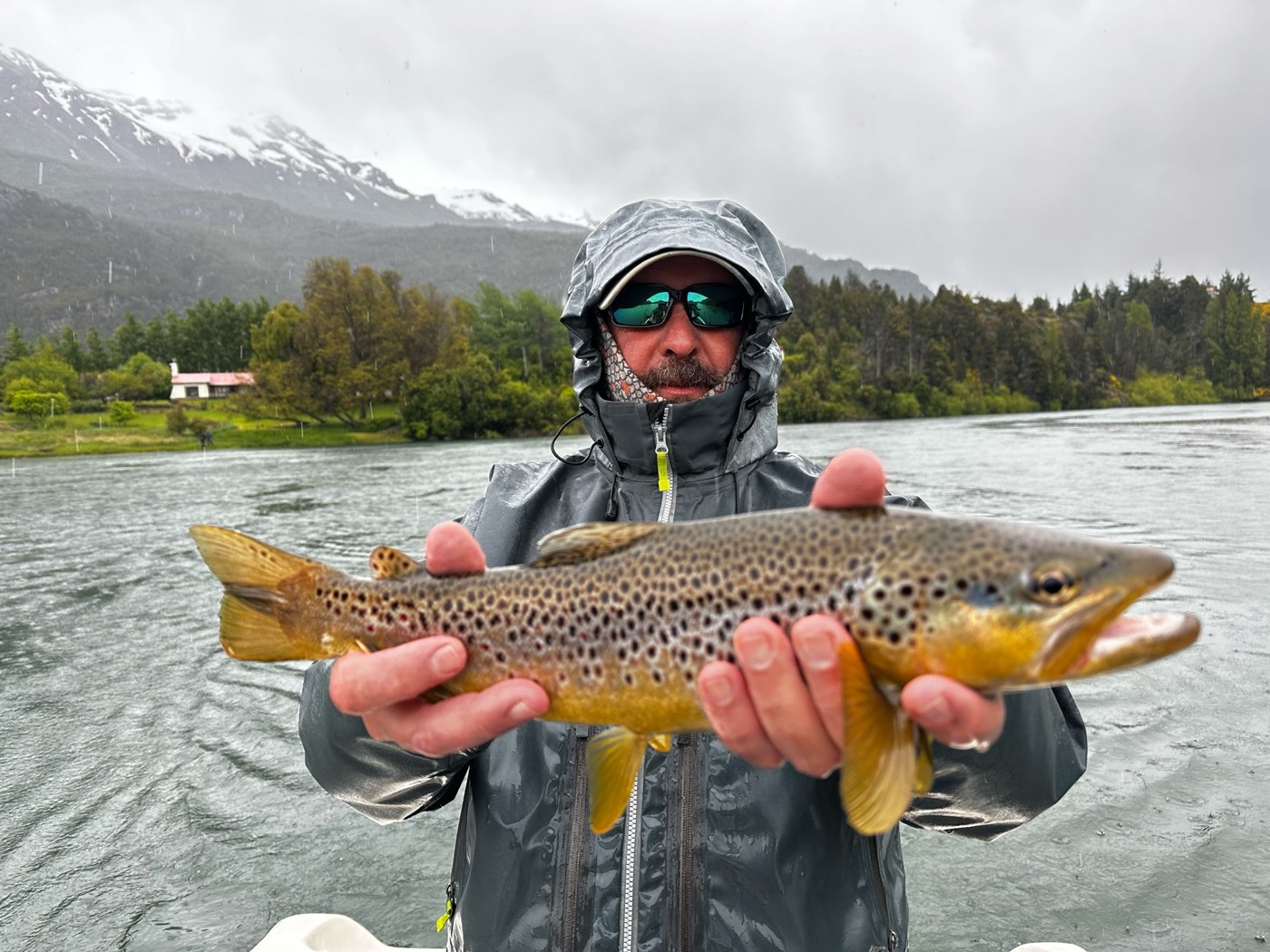 A guest holding a brown trout in the rain, snow-capped mountains and a lodge house across the water
