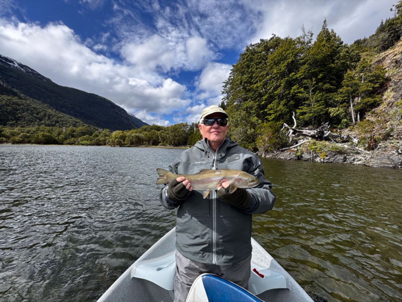 A guest holding a rainbow trout at the bow of a boat, a bright sky with scattered clouds over forested hills