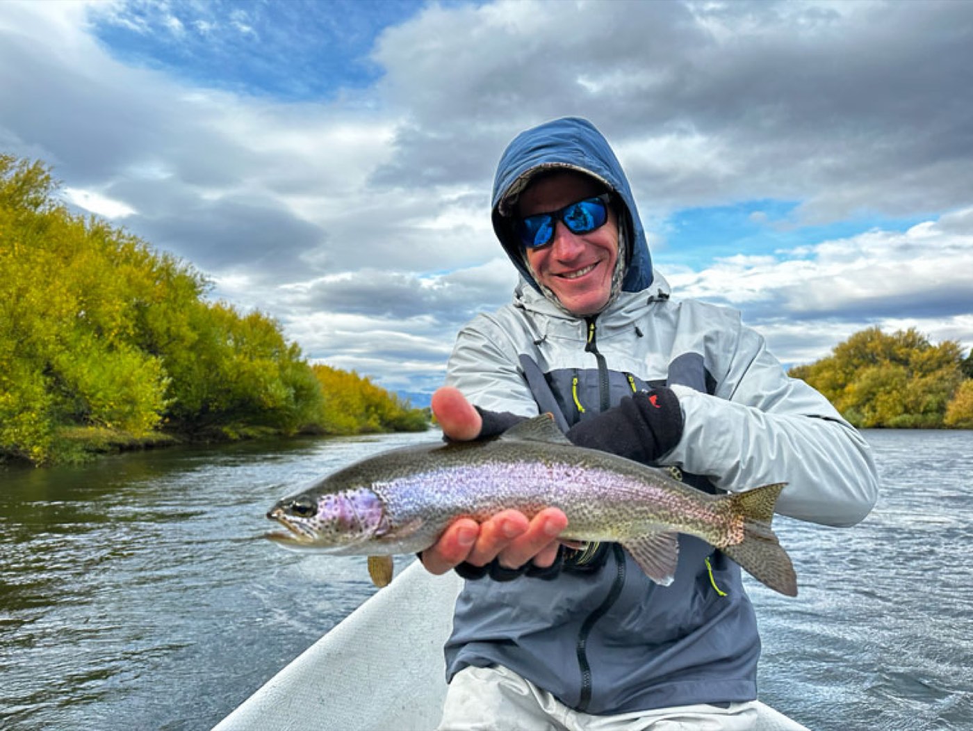 A guest holding a rainbow trout on a boat, golden autumn foliage lining the riverbank under an overcast sky