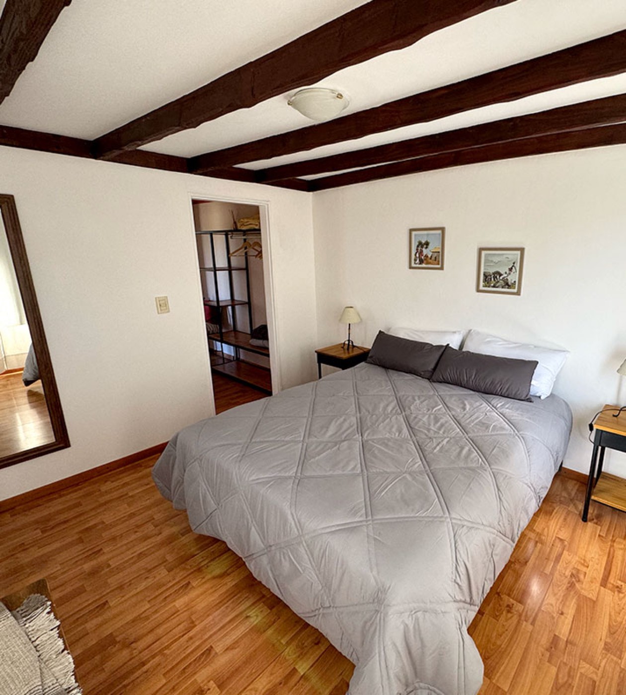A guest room with a grey comforter and pillows beneath an exposed wood-beam ceiling