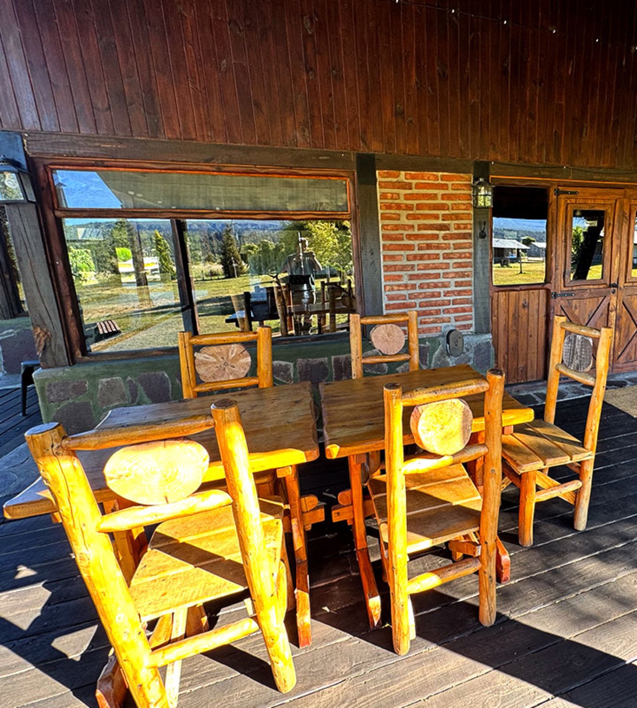 A covered outdoor dining area with a long wooden table and log-frame chairs, a wood-fired oven visible through the window