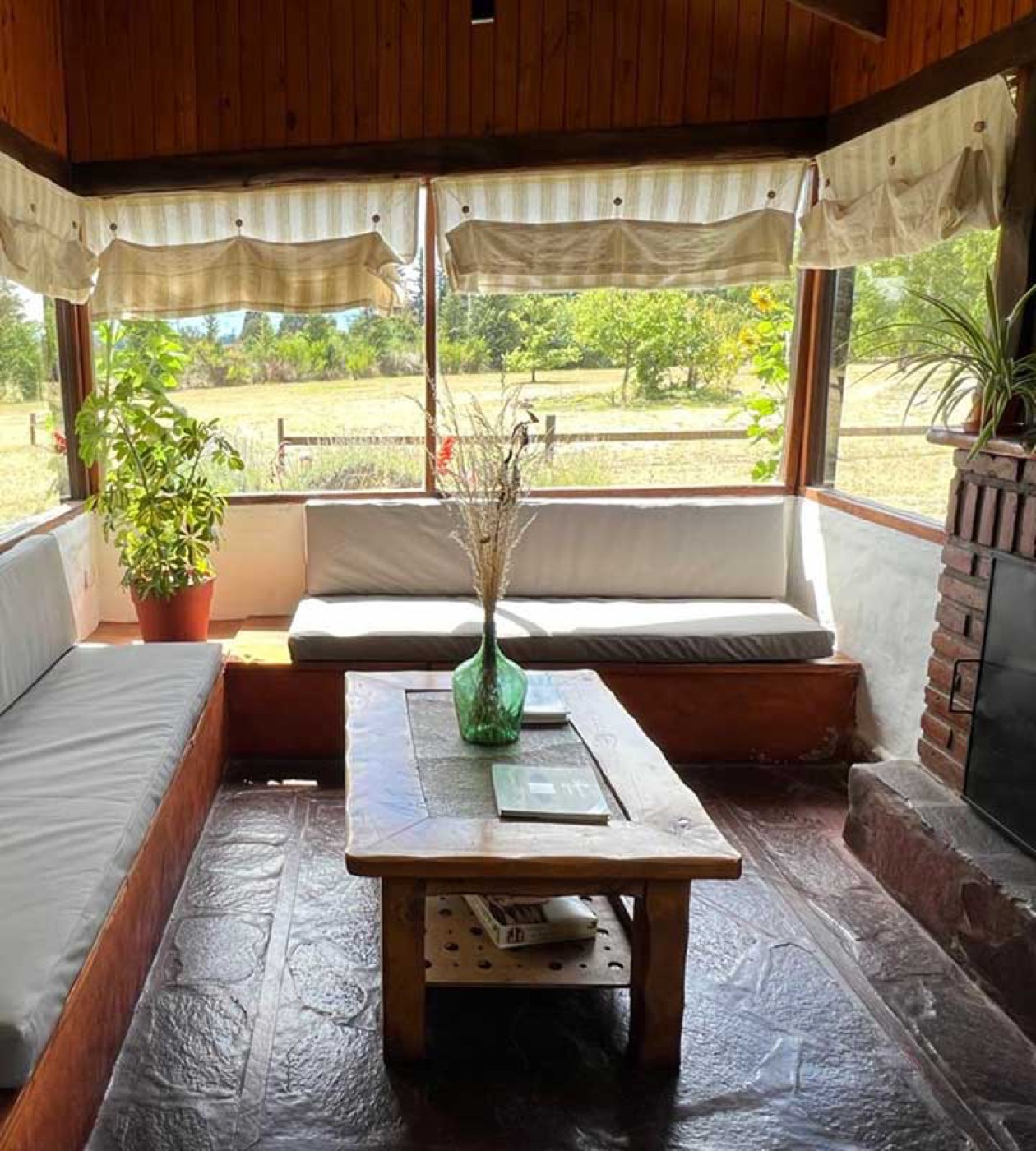 A sunroom lounge with built-in bench seating around a low wooden coffee table, overlooking open fields