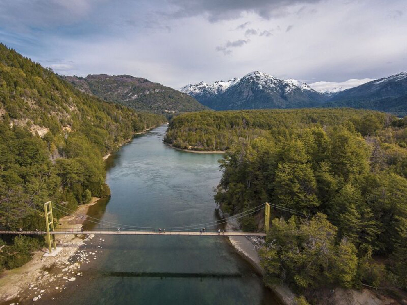 An aerial view of a suspension footbridge crossing the river, its teal water bending between forested slopes with snow-capped peaks behind