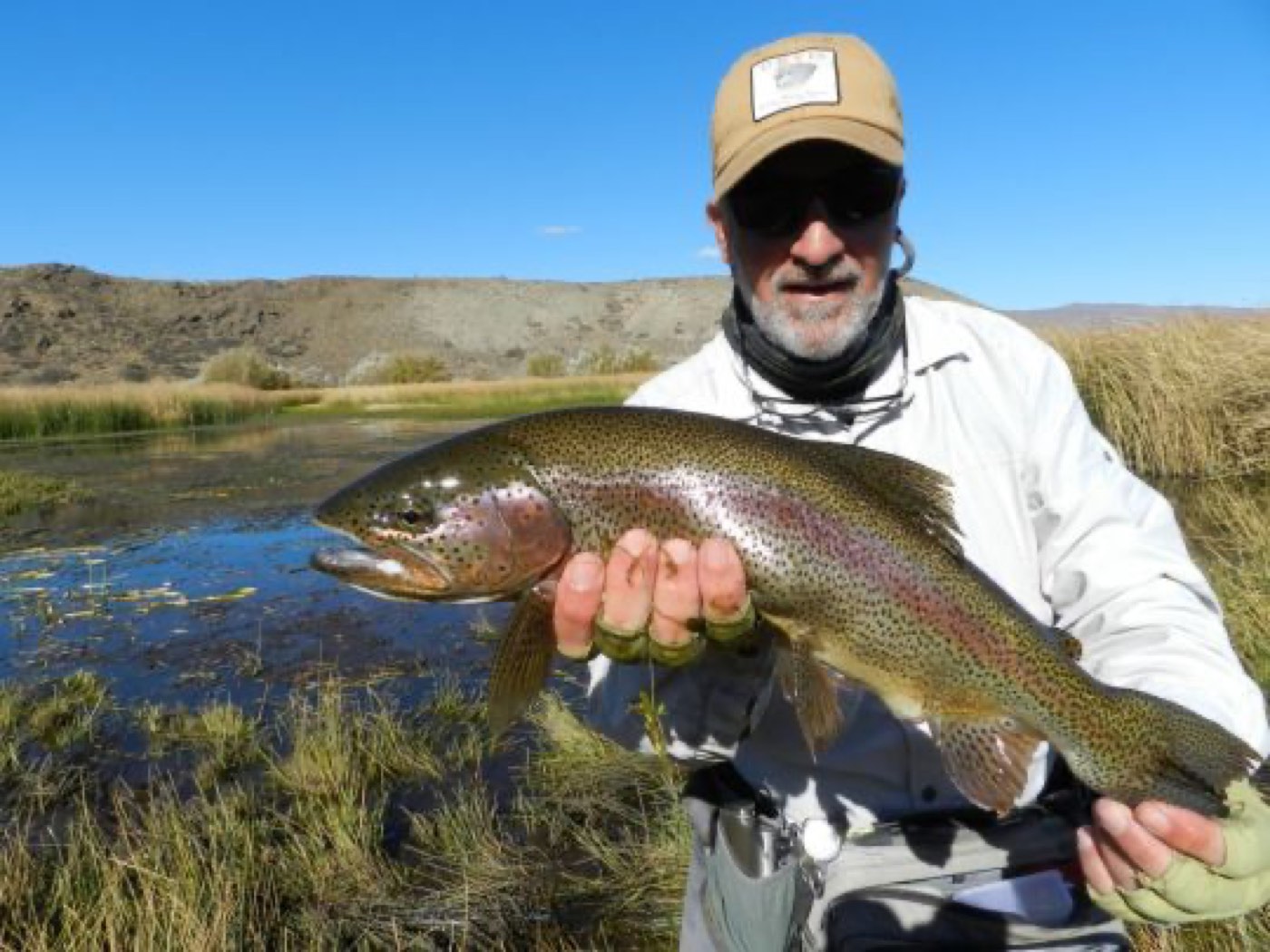 An angler holding a large rainbow trout beside a marshy pond, grassland and dry hills behind
