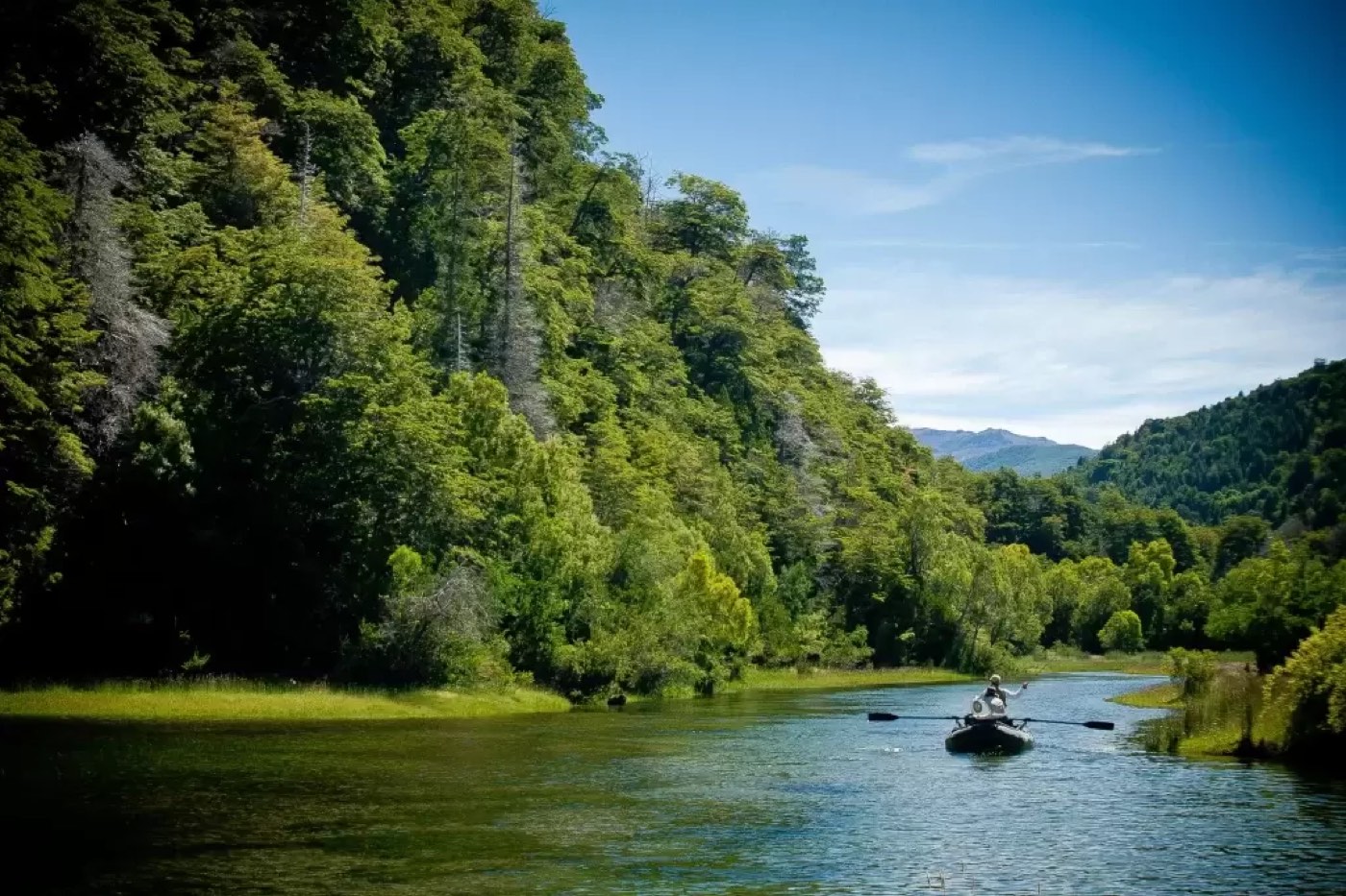 Someone rowing a drift boat down the river beneath a steep, forested hillside, mountains in the distance