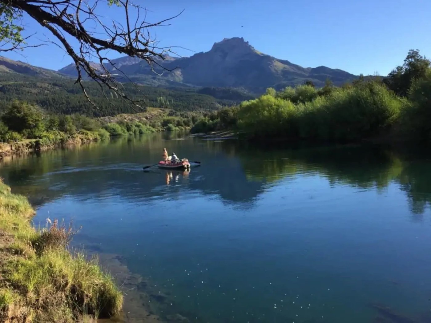 Two anglers floating the calm river in a small raft, a sharp mountain peak reflected in the water