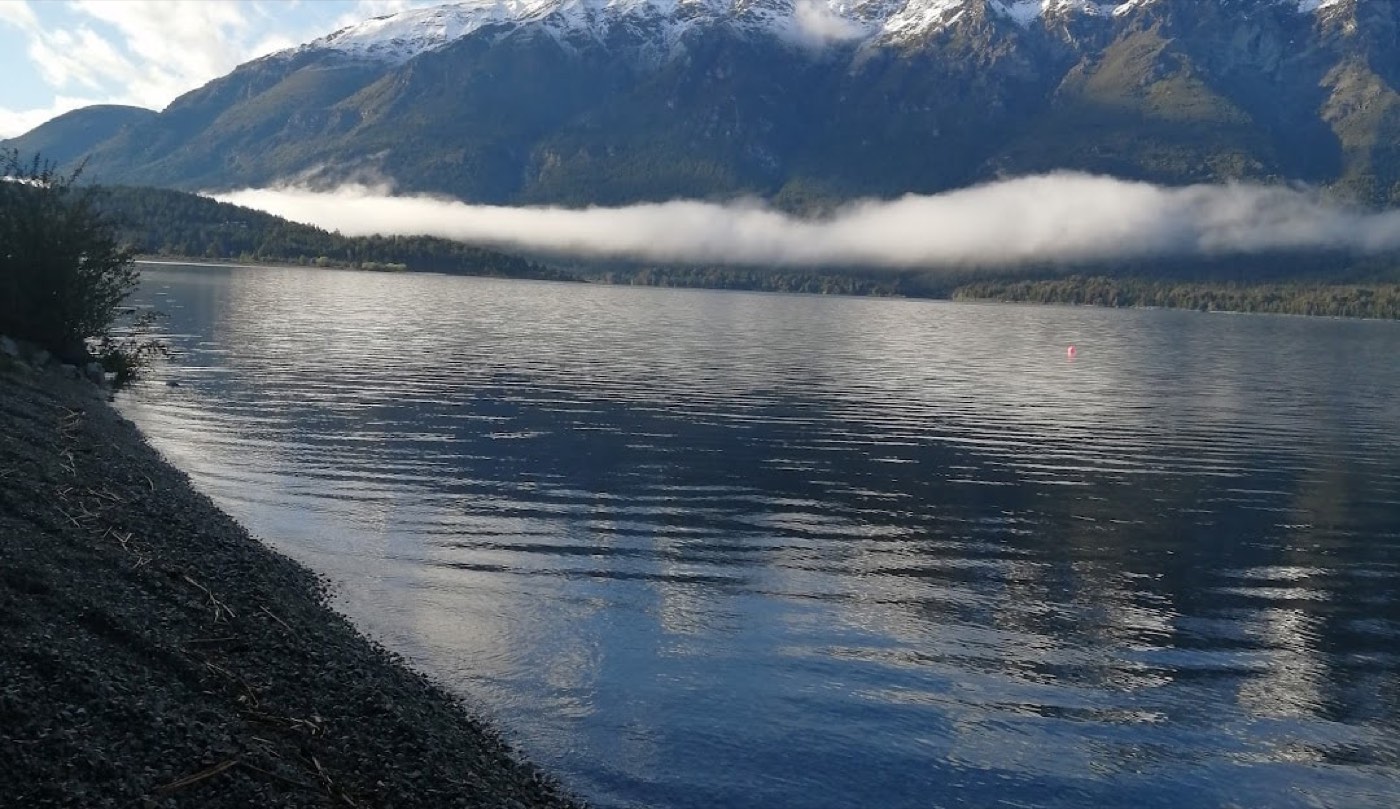 A low bank of cloud crossing snow-capped peaks above Lago Cholila's calm water, gravel shoreline in the foreground