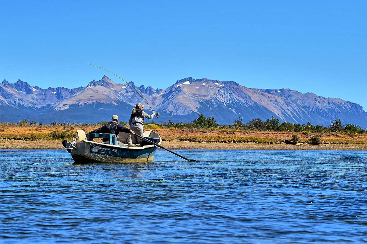 Two anglers in a drift boat on the Rio Corcovado, one standing and casting a fly rod toward a shoreline of golden grass, with jagged snow-flecked mountains and a clear blue sky behind