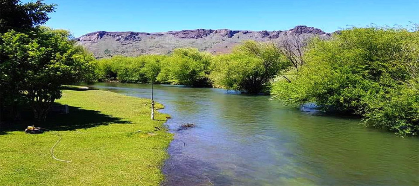 A calm stretch of river bordered by mown lawn on one bank and dense trees on the other, dry rocky hills behind