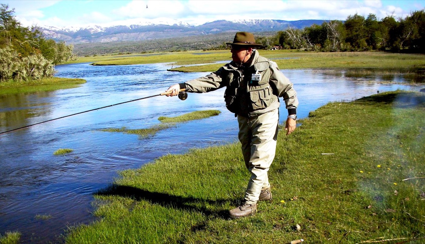 An angler in a wide-brimmed hat fly fishing from a wet grassy bank, marshland around him and snow-capped mountains beyond