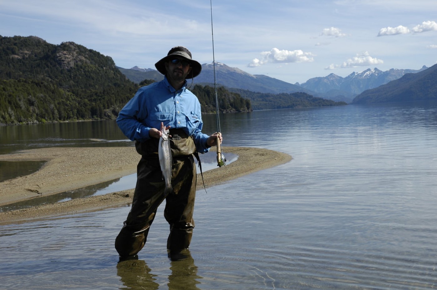 An angler wading in the shallows holding a trout and rod, a sandbar and calm lake stretching toward the mountains