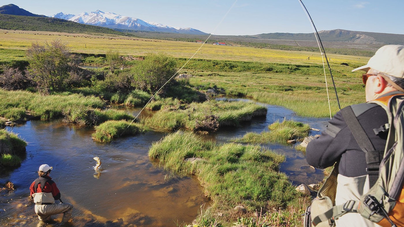 Two anglers fishing a grassy, spring-fed stream winding through wetland, snow-capped Andes on the horizon
