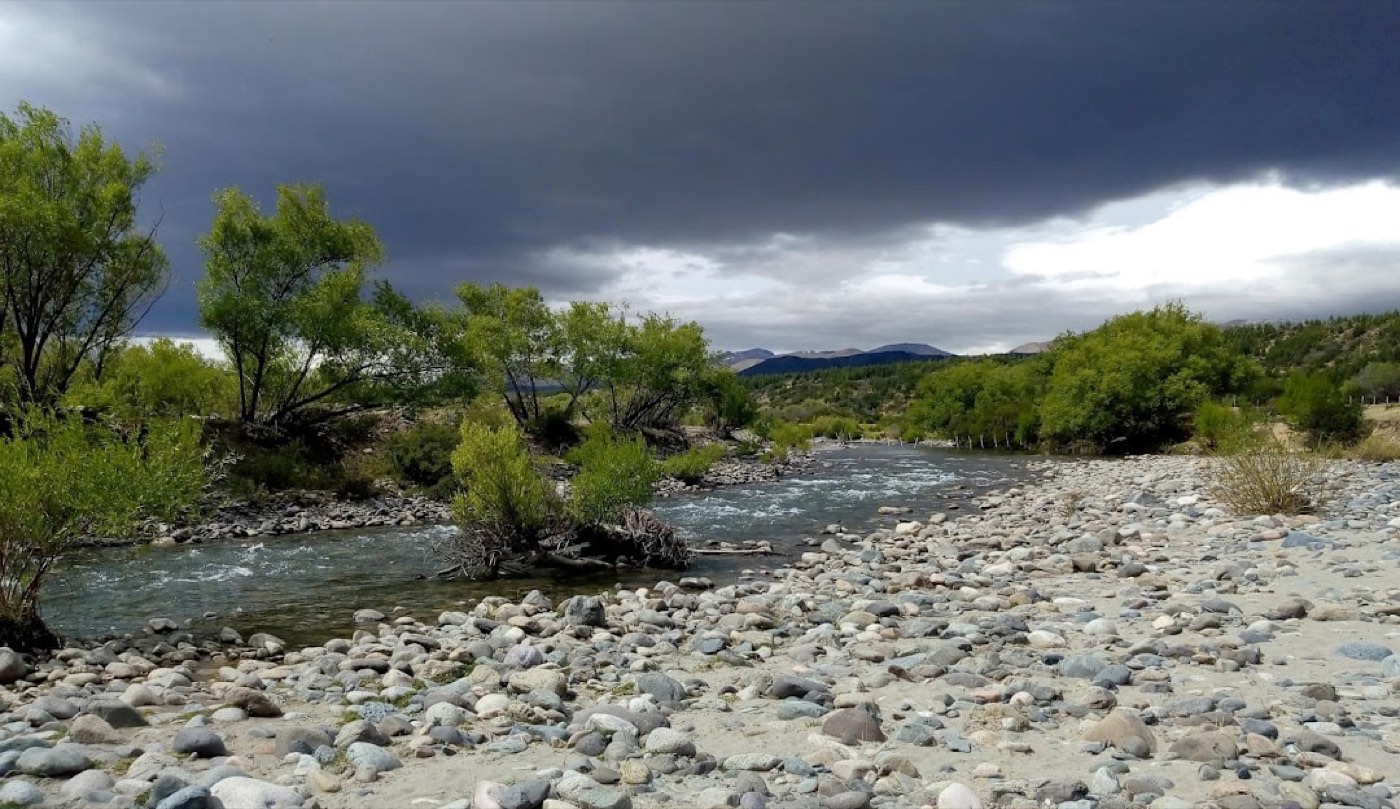 The river running low over gravel between willow-lined banks, dark storm clouds gathering overhead