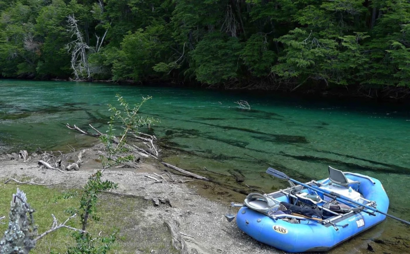 A blue raft beached on a gravel bank beside clear teal water, forest rising on the far shore