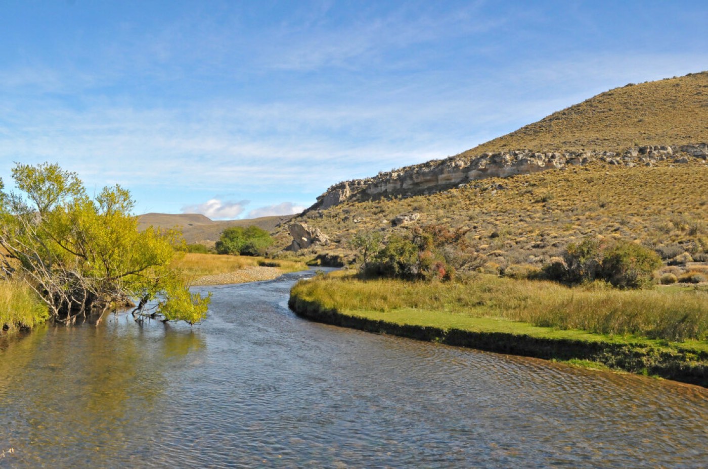 The stream bending past a grassy bank and a rocky, brush-covered hillside, distant peaks beyond