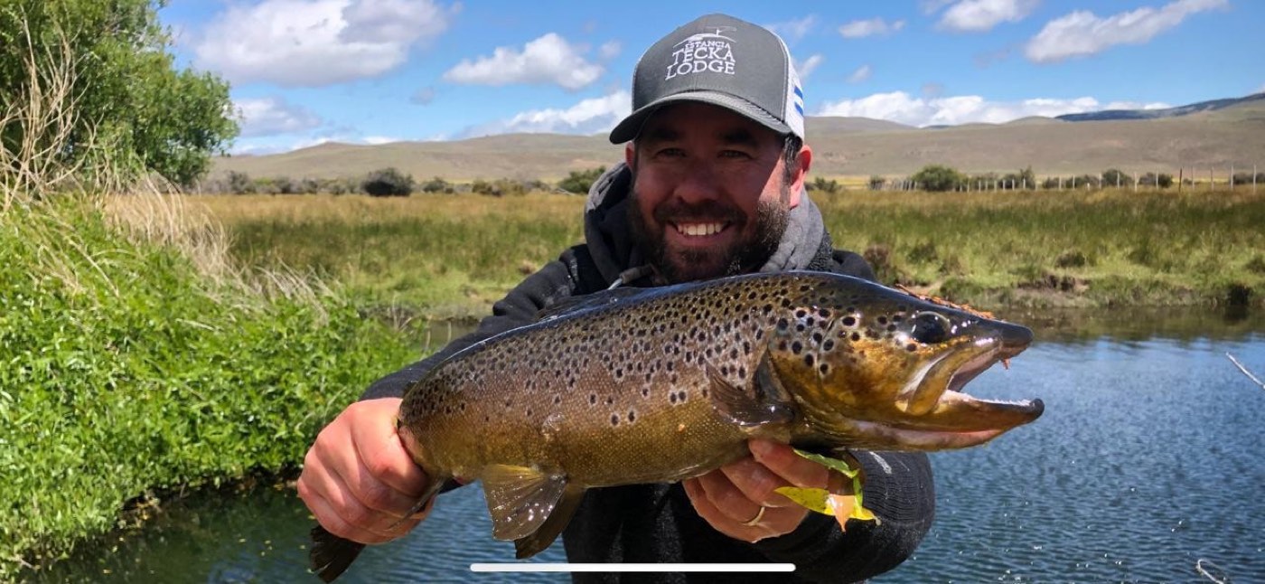 An angler in a ballcap holding a brown trout beside the water, grassland and hills behind