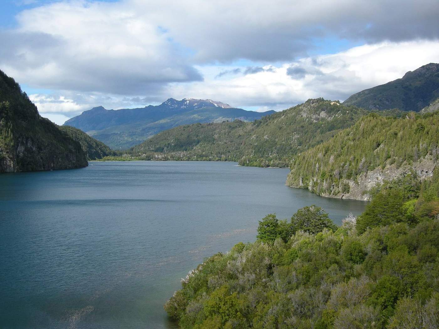 Lago Verde from above: a broad blue-green lake enclosed by steep forested hills and cliffs, with a snow-flecked peak and heavy clouds beyond